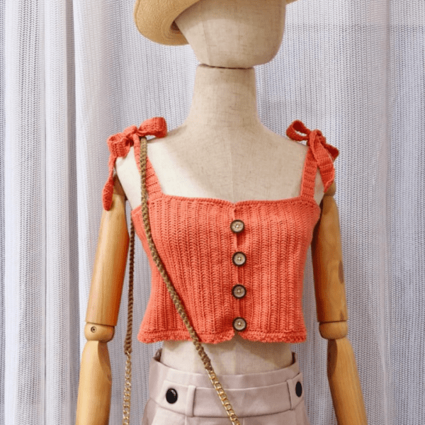 "Handmade coral-colored cotton top named 'Andrea Top' by Arty-Crafty by Vanessa, featuring adjustable shoulder ties and a square neckline, displayed on a mannequin with a straw hat and a small woven bag.