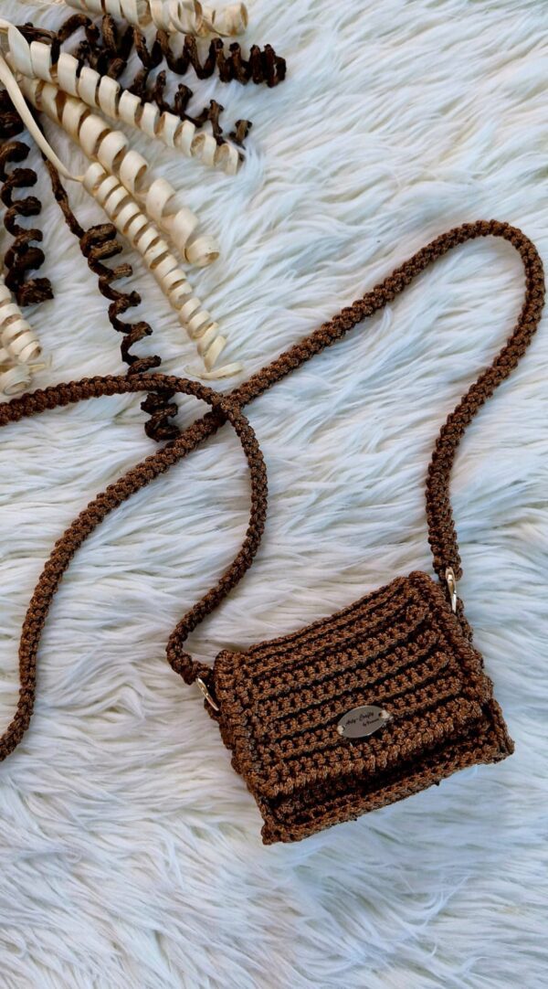 Handmade Nicky Purse in brown color, crafted from thick premium polyester yarn, featuring a compact design with a long strap, displayed on a white furry background.