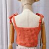 "Handmade coral-colored cotton top named 'Andrea Top' by Arty-Crafty by Vanessa, featuring adjustable shoulder ties and a square neckline, displayed on a mannequin with a straw hat and a small woven bag.