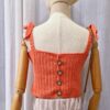 "Handmade coral-colored cotton top named 'Andrea Top' by Arty-Crafty by Vanessa, featuring adjustable shoulder ties and a square neckline, displayed on a mannequin with a straw hat and a small woven bag.