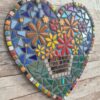 Handcrafted heart-shaped mosaic wall art featuring colorful glass tiles arranged in a vibrant floral pattern, with a basket of flowers at the center. The mosaic is designed with a mix of bright reds, yellows, oranges, blues, greens, and purples, creating a cheerful and lively decoration