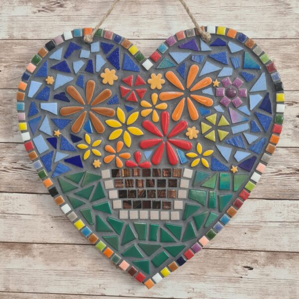 Handcrafted heart-shaped mosaic wall art featuring colorful glass tiles arranged in a vibrant floral pattern, with a basket of flowers at the center. The mosaic is designed with a mix of bright reds, yellows, oranges, blues, greens, and purples, creating a cheerful and lively decoration