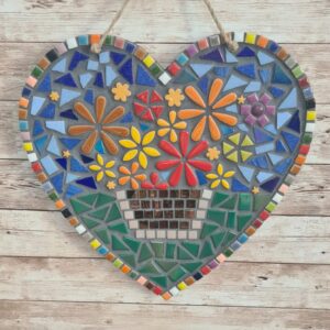 Handcrafted heart-shaped mosaic wall art featuring colorful glass tiles arranged in a vibrant floral pattern, with a basket of flowers at the center. The mosaic is designed with a mix of bright reds, yellows, oranges, blues, greens, and purples, creating a cheerful and lively decoration