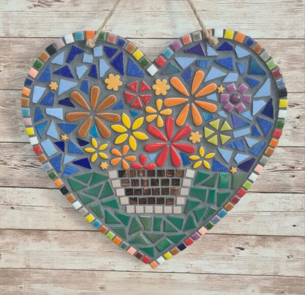 Handcrafted heart-shaped mosaic wall art featuring colorful glass tiles arranged in a vibrant floral pattern, with a basket of flowers at the center. The mosaic is designed with a mix of bright reds, yellows, oranges, blues, greens, and purples, creating a cheerful and lively decoration