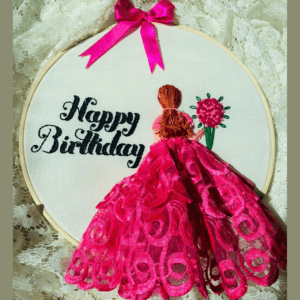 Hand-embroidered "Happy Birthday" wall art in a 10-inch wooden hoop featuring a pink lace 3D dress design and a girl holding a flower bouquet, created by Anu Paulson.