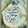 Alt Text: Hand-embroidered wall art featuring the phrase "The Secret Ingredient is Always Love," framed in a 10-inch wooden hoop with colorful cooking-themed designs, created by Anu Paulson.