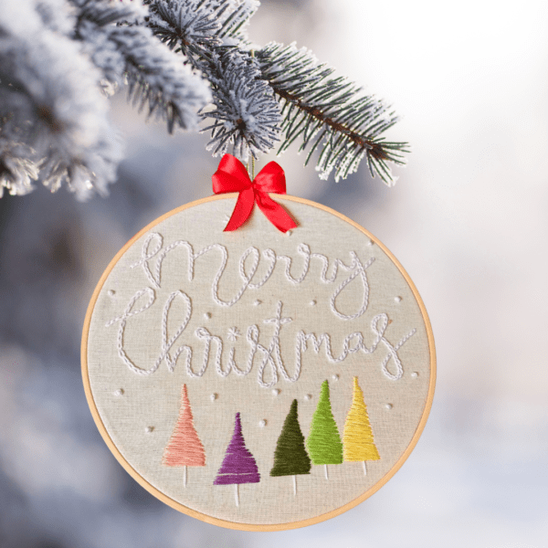 Hand-embroidered "Merry Christmas" wall art featuring colorful Christmas trees, framed in an 8-inch wooden hoop with a red ribbon, created by Anu Paulson.