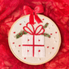 Hand-embroidered wall art featuring a festive Christmas gift, framed in an 8-inch wooden hoop with a red bow, created by Anu Paulson.