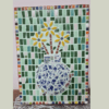 A colorful mosaic artwork featuring a vase filled with yellow flowers. The vase is composed of blue and white ceramic pieces, arranged on a background of green, pink, and white tiles. The border of the mosaic is framed with small, multicolored tiles, adding vibrant contrast to the piece.