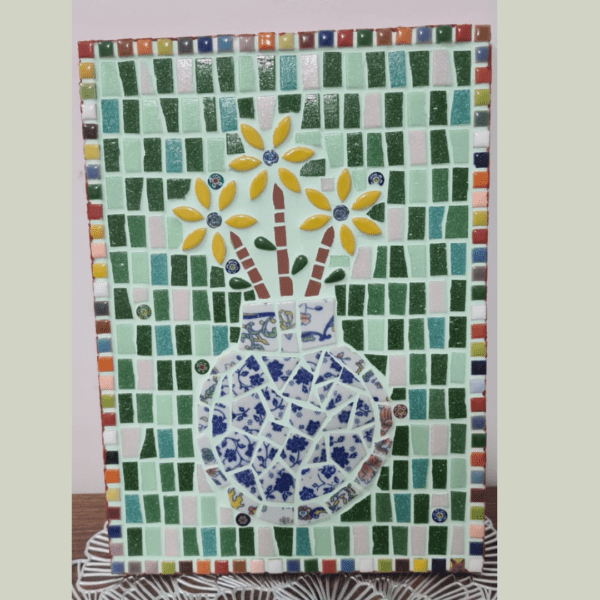 A colorful mosaic artwork featuring a vase filled with yellow flowers. The vase is composed of blue and white ceramic pieces, arranged on a background of green, pink, and white tiles. The border of the mosaic is framed with small, multicolored tiles, adding vibrant contrast to the piece.