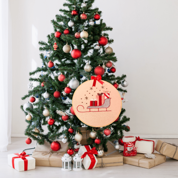 Hand-embroidered wall art featuring a Christmas sleigh filled with gifts, framed in an 8-inch wooden hoop with a festive red bow, created by Anu Paulson.