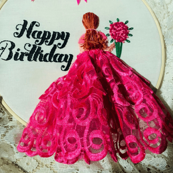 Hand-embroidered "Happy Birthday" wall art in a 10-inch wooden hoop featuring a pink lace 3D dress design and a girl holding a flower bouquet, created by Anu Paulson.