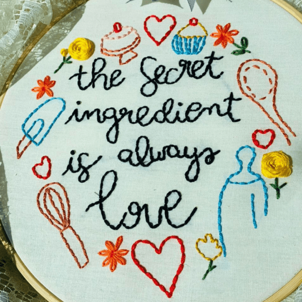 Alt Text: Hand-embroidered wall art featuring the phrase "The Secret Ingredient is Always Love," framed in a 10-inch wooden hoop with colorful cooking-themed designs, created by Anu Paulson.