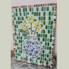 A colorful mosaic artwork featuring a vase filled with yellow flowers. The vase is composed of blue and white ceramic pieces, arranged on a background of green, pink, and white tiles. The border of the mosaic is framed with small, multicolored tiles, adding vibrant contrast to the piece.