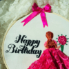 Hand-embroidered "Happy Birthday" wall art in a 10-inch wooden hoop featuring a pink lace 3D dress design and a girl holding a flower bouquet, created by Anu Paulson.