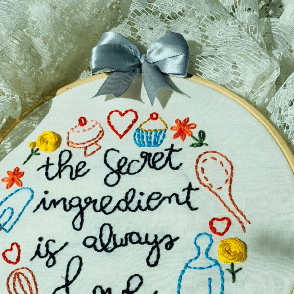 Alt Text: Hand-embroidered wall art featuring the phrase "The Secret Ingredient is Always Love," framed in a 10-inch wooden hoop with colorful cooking-themed designs, created by Anu Paulson.