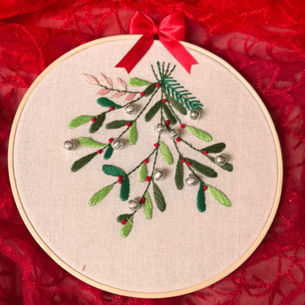 Hand-embroidered Christmas mistletoe wall art featuring green mistletoe leaves, red berries, and silver bells, framed in an 8-inch wooden hoop with a red ribbon, created by Anu Paulson.