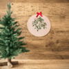 Hand-embroidered Christmas mistletoe wall art featuring green mistletoe leaves, red berries, and silver bells, framed in an 8-inch wooden hoop with a red ribbon, created by Anu Paulson.