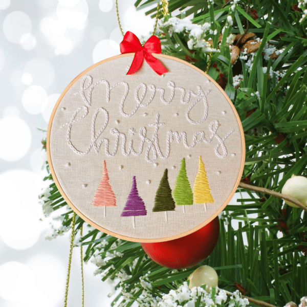 Hand-embroidered "Merry Christmas" wall art featuring colorful Christmas trees, framed in an 8-inch wooden hoop with a red ribbon, created by Anu Paulson.