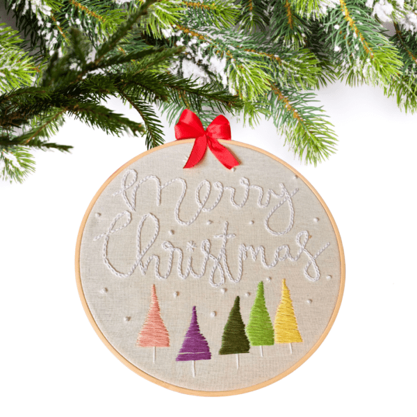 Hand-embroidered "Merry Christmas" wall art featuring colorful Christmas trees, framed in an 8-inch wooden hoop with a red ribbon, created by Anu Paulson.