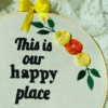 Hand-embroidered wall art in a 10-inch wooden hoop with the message "This is Our Happy Place," decorated with yellow and orange floral details, created by Anu Paulson.