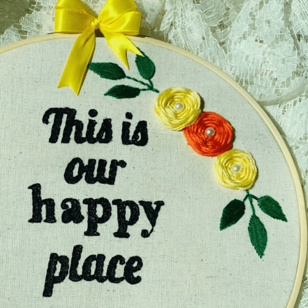 Hand-embroidered wall art in a 10-inch wooden hoop with the message "This is Our Happy Place," decorated with yellow and orange floral details, created by Anu Paulson.