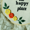 Hand-embroidered wall art in a 10-inch wooden hoop with the message "This is Our Happy Place," decorated with yellow and orange floral details, created by Anu Paulson.