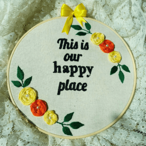 Hand-embroidered wall art in a 10-inch wooden hoop with the message "This is Our Happy Place," decorated with yellow and orange floral details, created by Anu Paulson.