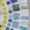 A round mosaic mirror featuring a pattern of rectangular ceramic tiles in varying shades of blue, white, and yellow, arranged in concentric circles around the central mirror. The design is mounted on an MDF base, creating a vibrant, artistic wall décor piece.