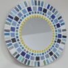 A round mosaic mirror featuring a pattern of rectangular ceramic tiles in varying shades of blue, white, and yellow, arranged in concentric circles around the central mirror. The design is mounted on an MDF base, creating a vibrant, artistic wall décor piece.