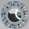 A round mosaic mirror featuring a pattern of rectangular ceramic tiles in varying shades of blue, white, and yellow, arranged in concentric circles around the central mirror. The design is mounted on an MDF base, creating a vibrant, artistic wall décor piece.