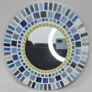 A round mosaic mirror featuring a pattern of rectangular ceramic tiles in varying shades of blue, white, and yellow, arranged in concentric circles around the central mirror. The design is mounted on an MDF base, creating a vibrant, artistic wall décor piece.