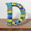 A handcrafted mosaic letter "D" featuring vibrant glass tiles in shades of blue, yellow, green, and purple, fixed on an MDF base. This colorful letter is perfect for home décor, kids' rooms, office spaces, or creating custom nameplates.