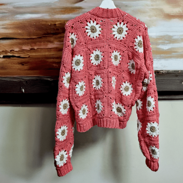 Handmade crochet Daisy Mae Cardigan in a warm coral color, adorned with white and beige daisy motifs. The cardigan features a buttoned front and a relaxed fit, perfect for layering on cool days.