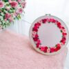 Hand-embroidered floral wreath wall art featuring pink, red, and blush roses with green leaves, framed in a 10-inch wooden hoop, created by Anu Paulson.