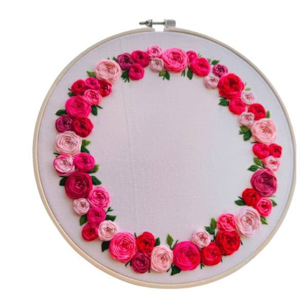 Hand-embroidered floral wreath wall art featuring pink, red, and blush roses with green leaves, framed in a 10-inch wooden hoop, created by Anu Paulson.