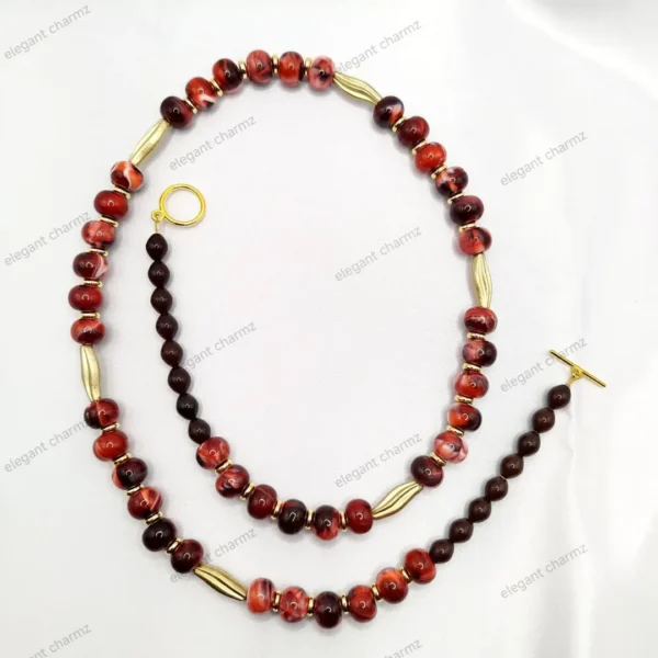 Handmade ceramic bead necklace and bracelet set in red and black with gold accents, laid on a white fabric background.