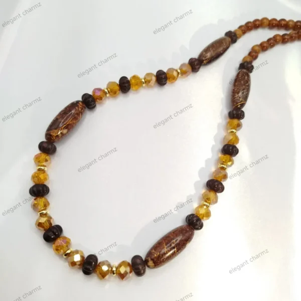 Handmade beaded necklace in brown and amber tones with ceramic and crystal beads, laid on a white fabric background.