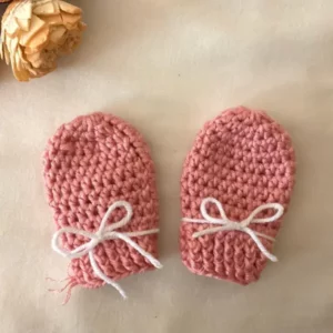 Pink handmade crochet baby mittens with white bow ties, displayed on a light background.