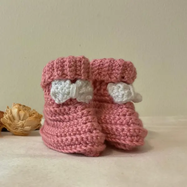 Handmade crochet baby booties in pink with white bow detail, standing upright on a light background.