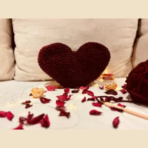 Handmade burgundy crochet heart cushion made with soft textured chenille yarn displayed on a sofa.